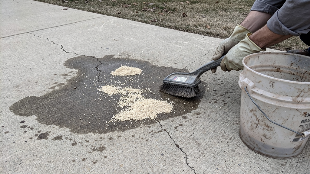 Oil stain on a concrete driveway beside absorbent material and a stiff brush